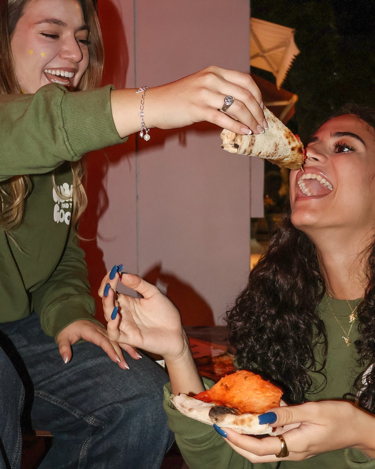 One friend feeding another a folded slice of pizza, both laughing