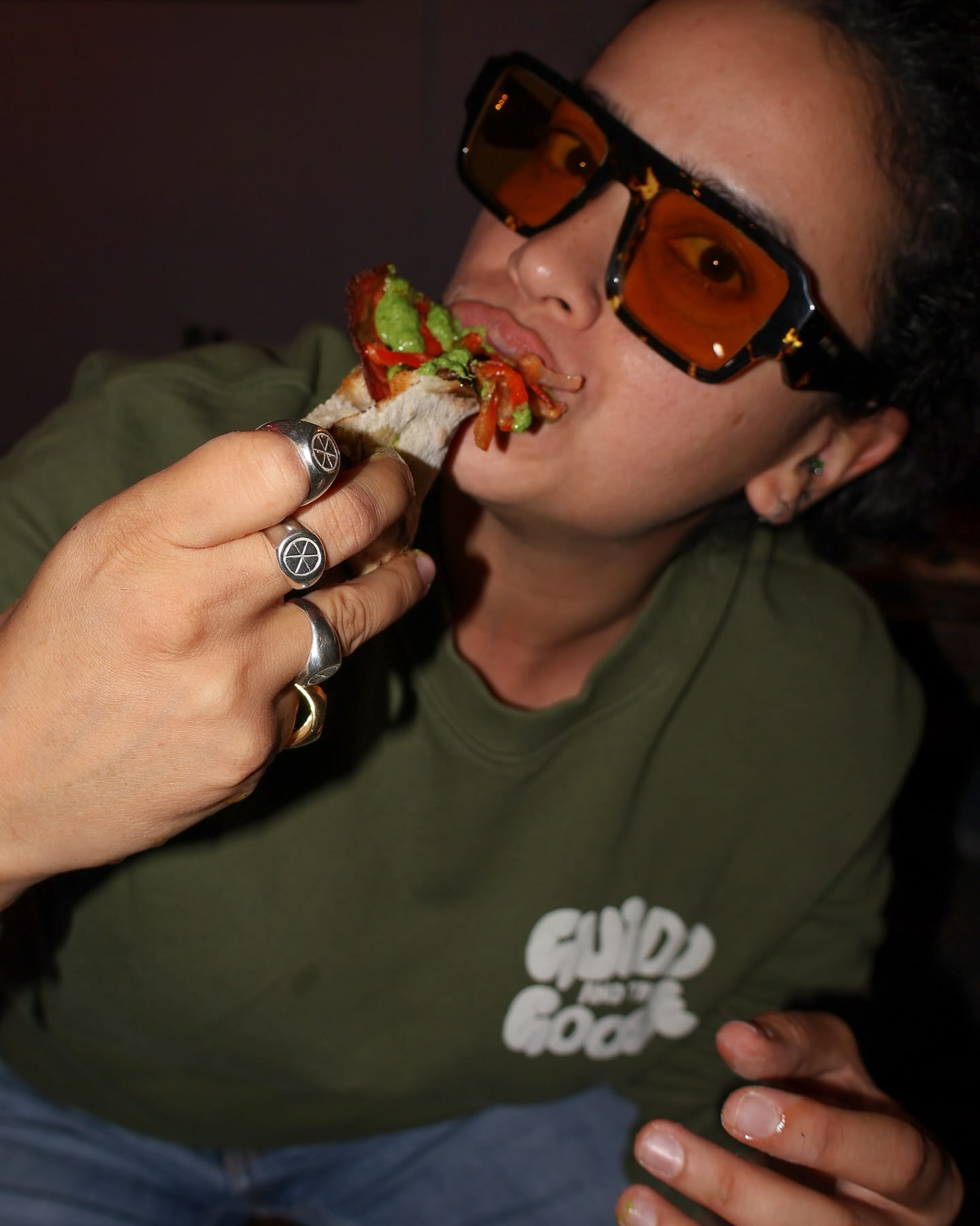 Eating a loaded slice while wearing signet rings and orange sunglasses