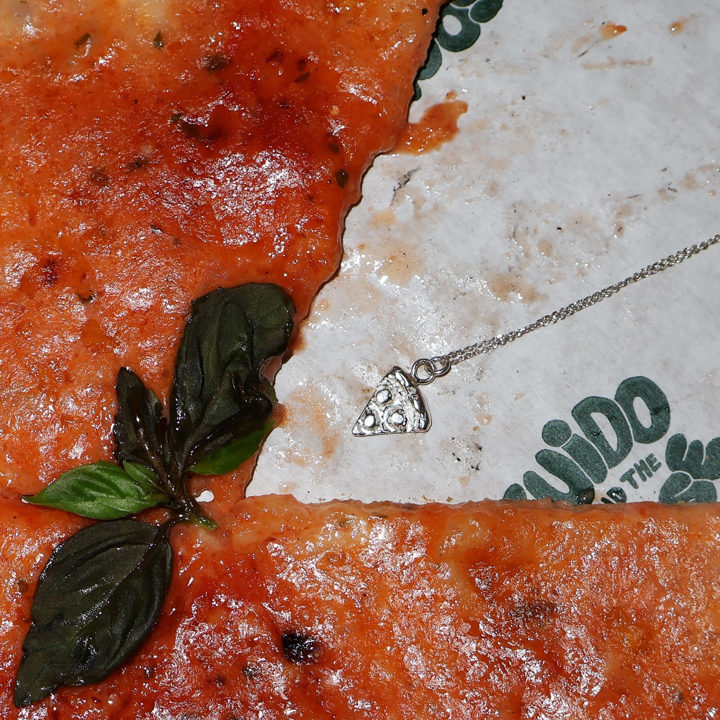 Close-up of pizza with basil and a silver pizza-slice necklace on Guido & The Goose paper